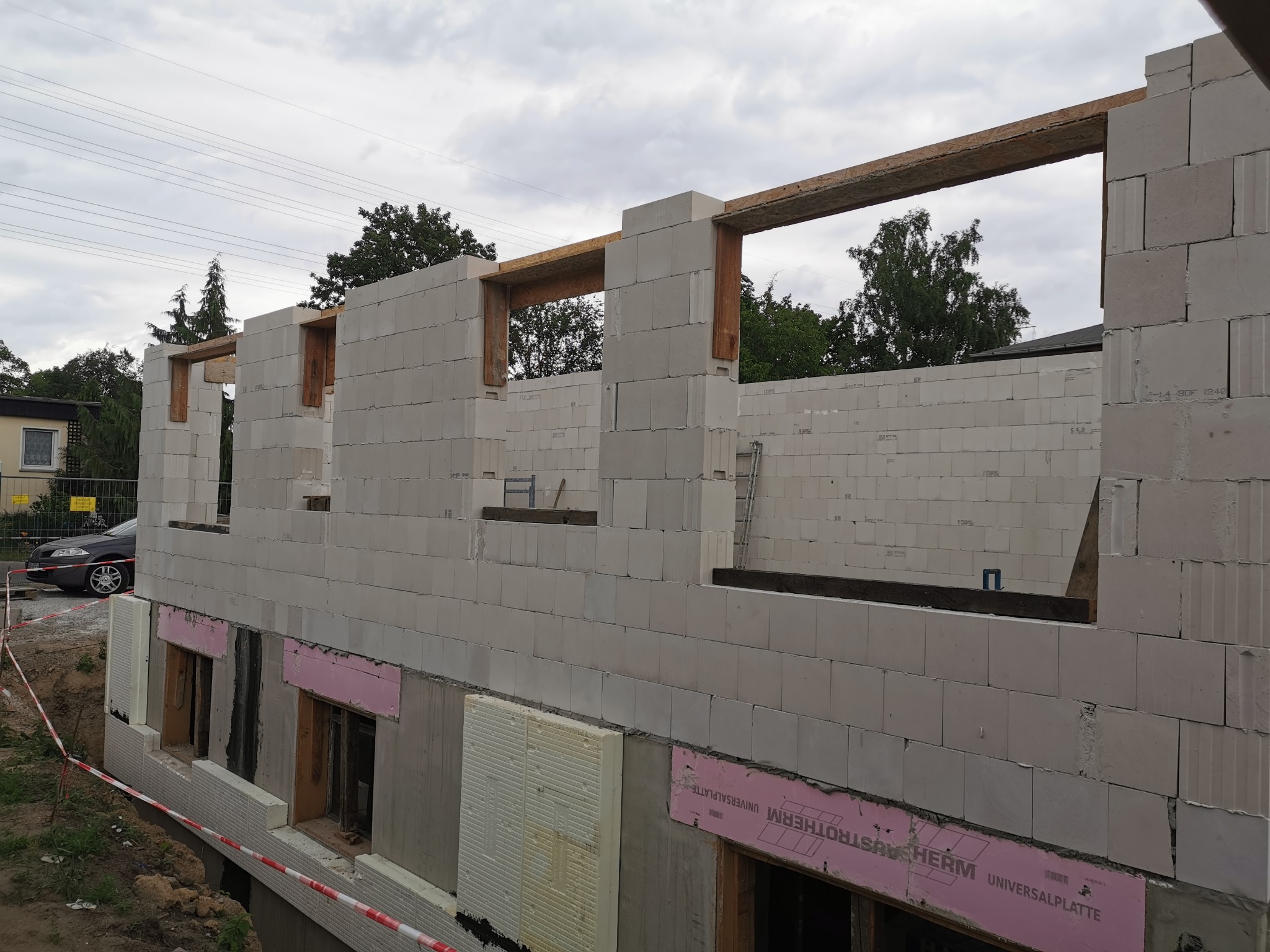 Wznoszenie ścian zewnętrznych budynku z bloczków betonowych, widoczne otwory okienne z drewnianymi nadprożami, ocieplenie fundamentów styropianem, teren budowy zabezpieczony taśmą.