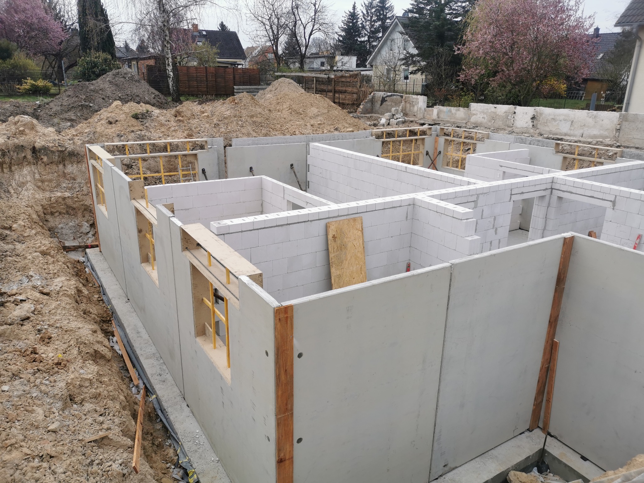 Wznoszenie fundamentów budynku z betonu i bloczków, widoczne szalunki okienne, ziemia wokół fundamentów, drzewa w tle.