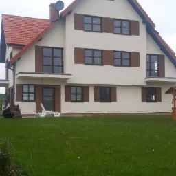 Dwupiętrowy dom jednorodzinny z czerwoną dachówką i brązowymi okiennicami, widziany z poziomu trawnika.