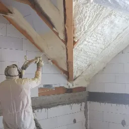Pracownik w kombinezonie ochronnym aplikuje białą izolację natryskową na drewnianą konstrukcję dachu w surowym wnętrzu budynku z białych bloczków.