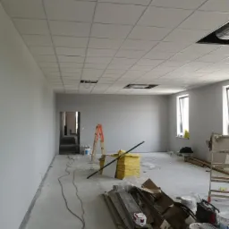 Montaż sufitu podwieszanego kasetonowego w szarym biurze. Widoczne płyty sufitowe, drabina, materiały izolacyjne i narzędzia na podłodze. Otwory na oświetlenie w suficie.