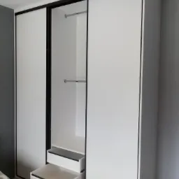 Biała szafa przesuwna z czarnymi obramowaniami, częściowo otwarta z wysuniętymi szufladami i widocznymi drążkami na ubrania.