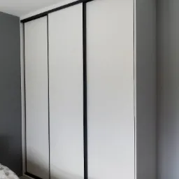 Biała szafa wnękowa z trzema przesuwanymi drzwiami z czarnymi obramowaniami, widoczna w rogu sypialni z szarą ścianą i częściowo widoczną pościelą w delikatne wzory.