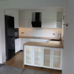 Nowoczesna, biała kuchnia z drewnianym blatem i czarnymi akcentami, w trakcie montażu, z widoczną folią ochronną na podłodze.