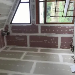 Pokój na poddaszu w trakcie prac wykończeniowych: widoczne płyty gipsowo-kartonowe z zaszpachlowanymi łączeniami, przygotowane do dalszych prac, okno dachowe w kształcie trójkąta, oraz rusztowanie...