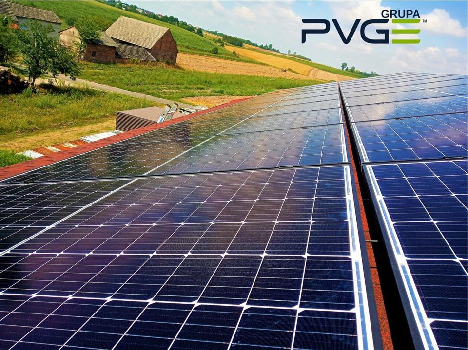Rząd paneli słonecznych na dachu budynku wiejskiego z logo firmy PVG w prawym górnym rogu, w tle pola i zabudowania.