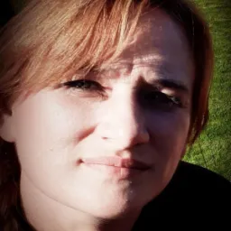 Portret kobiety z rudymi włosami na tle zielonej trawy, oświetlonej słońcem, z widocznymi cieniami na twarzy. Ujęcie z bliska, skupiające się na mimice.