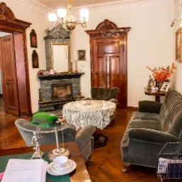 Wnętrze gabinetu z drewnianymi elementami, kominkiem, antykami i zielonym biurkiem z lampą. Na biurku leżą dokumenty i filiżanka.