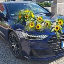Granatowy samochód marki Audi udekorowany bukietami słoneczników i białych kwiatów, stojący na brukowanym placu przed pomnikiem.