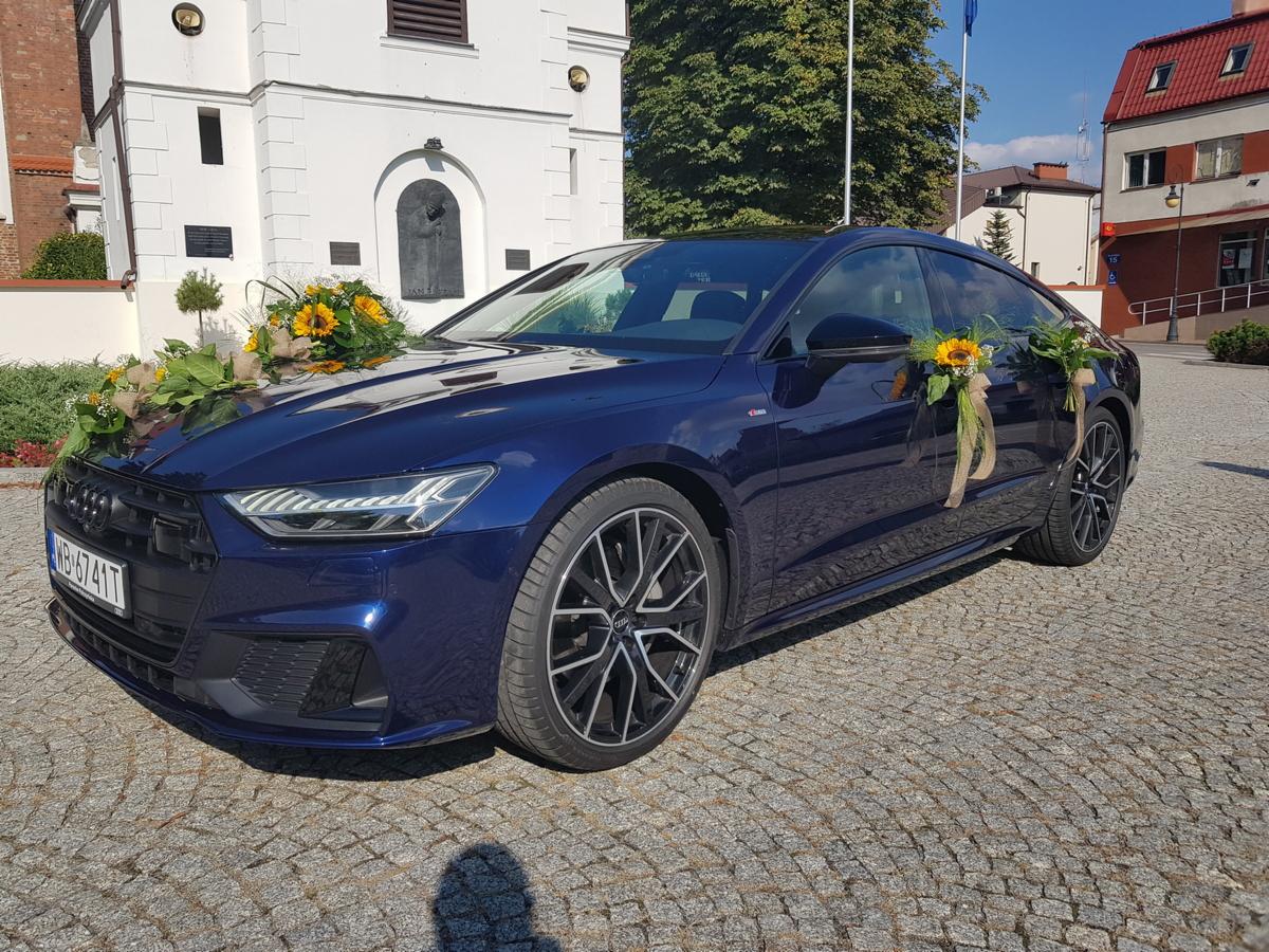 Niebieski samochód Audi A7 udekorowany słonecznikami i jutowymi wstążkami, zaparkowany na brukowanej ulicy przed białym budynkiem z elementami architektury sakralnej.