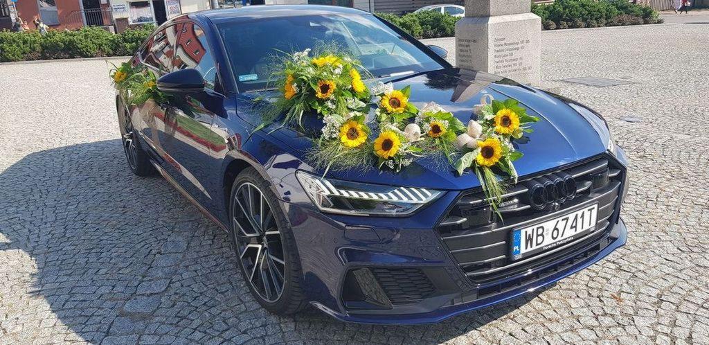 Granatowy samochód marki Audi udekorowany bukietami słoneczników i białych kwiatów, stojący na brukowanym placu przed pomnikiem.