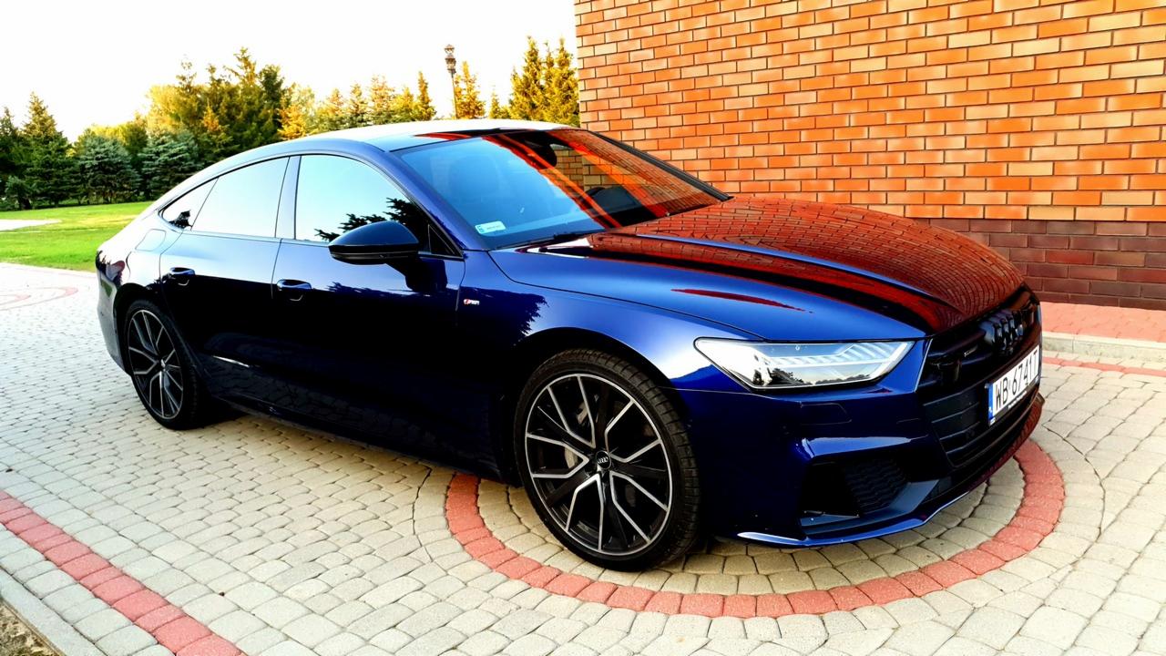 Błyszczący, granatowy Audi A7 zaparkowany na kostce brukowej z czerwoną obwódką, na tle ceglanego budynku i zieleni.