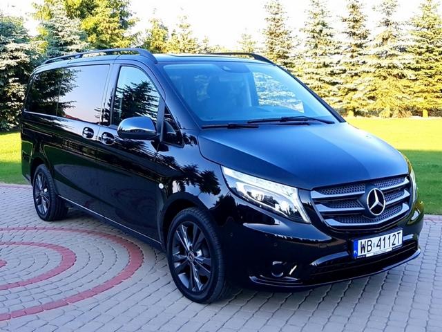 Czarny, luksusowy van Mercedes V-klasy z relingami dachowymi, zaparkowany na kostce brukowej przed zielonymi drzewami.