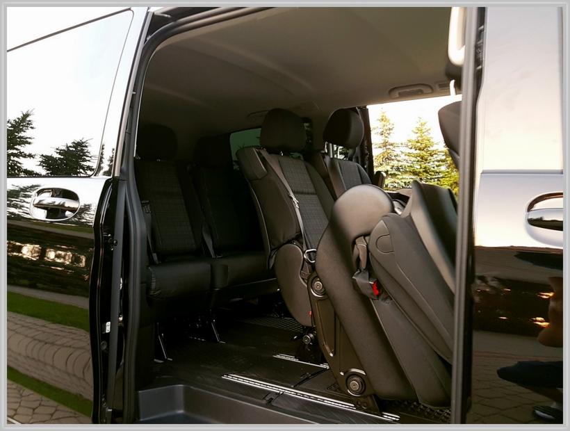 Wnętrze czarnego vana z otwartymi drzwiami, widoczne trzy rzędy foteli z szarej tapicerki w kratę, drzewa iglaste odbijające się w lakierze karoserii.