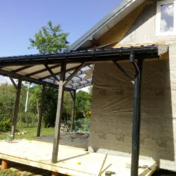 Drewniany taras w trakcie budowy z widocznymi elementami konstrukcyjnymi, czarną rynną i fragmentem elewacji budynku w tle.