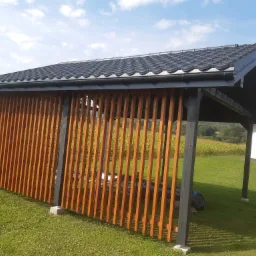 Nowoczesna altana z drewnianymi listwami, czarną konstrukcją i dachówką, widok na zielony trawnik i pole kukurydzy w tle.