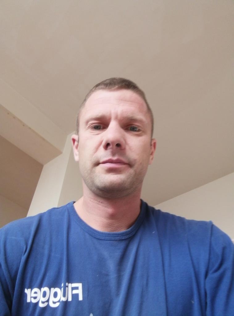 Mężczyzna w niebieskiej koszulce z krótkim rękawem, patrzący w obiektyw, z widocznym fragmentem sufitu w tle. Ujęcie z dołu.