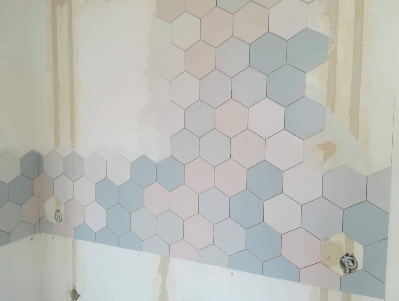 Ściana w trakcie układania heksagonalnych płytek w pastelowych odcieniach błękitu, różu i bieli, widoczne puste przestrzenie po płytkach wokół gniazdek elektrycznych, ślady po taśmie malarskiej...