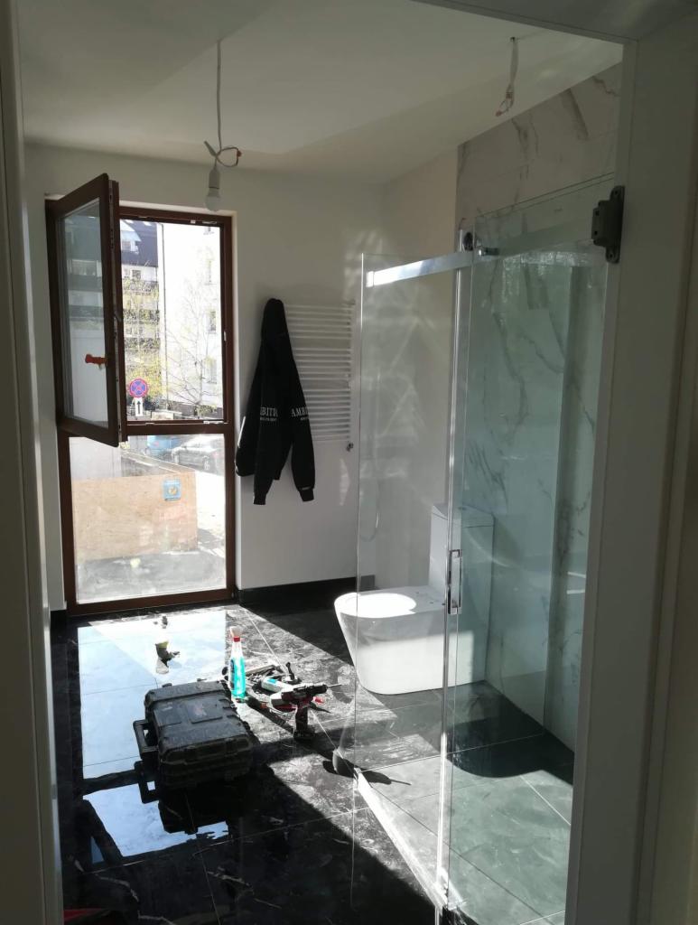 Nowoczesna łazienka w trakcie remontu: czarne, błyszczące płytki podłogowe, częściowo zamontowana szklana kabina prysznicowa, narzędzia i akcesoria na podłodze, otwarte okno z widokiem na budynek.