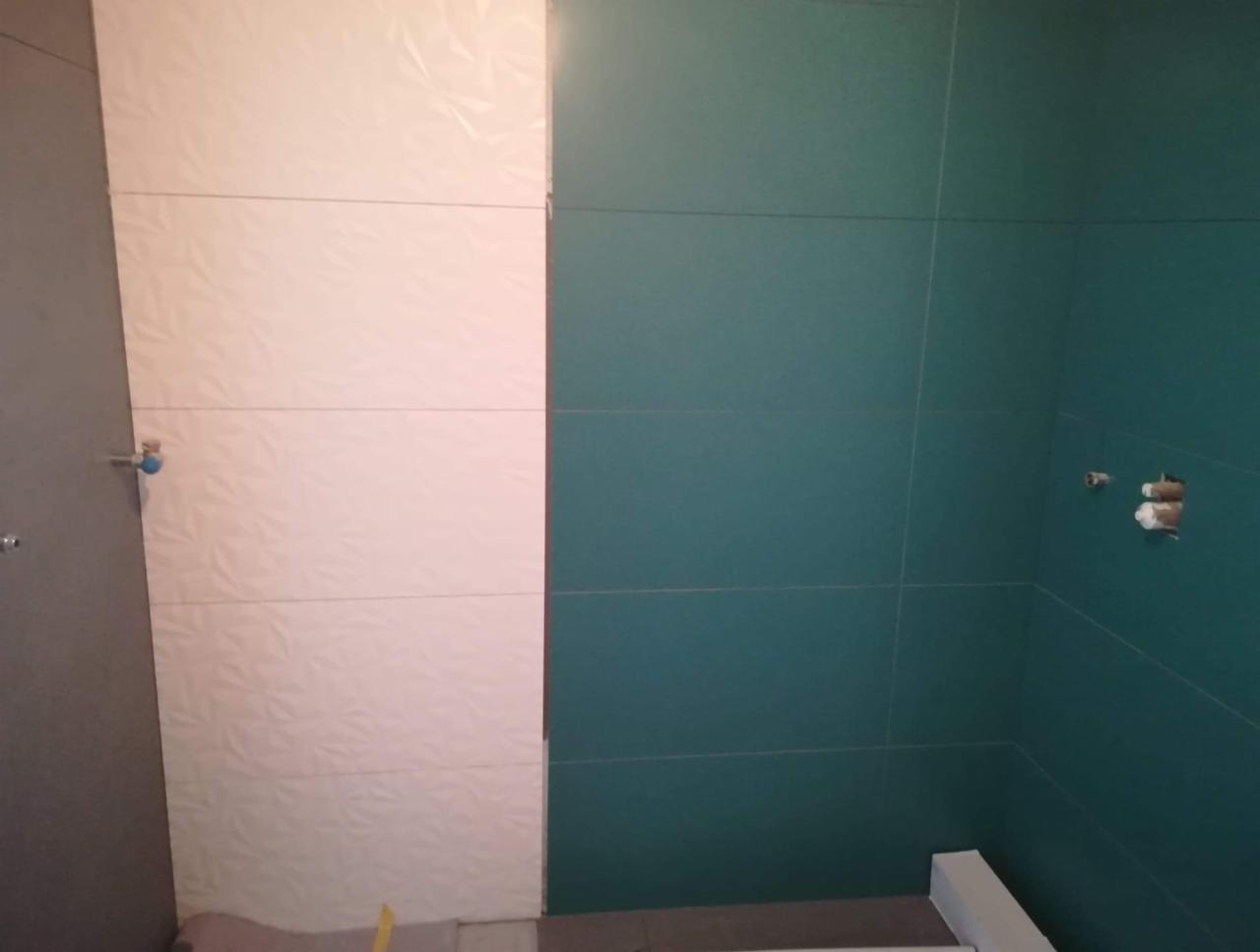 Ściana łazienki wyłożona białymi, trójwymiarowymi płytkami oraz turkusowymi płytkami, widoczne przygotowane przyłącza wodne.