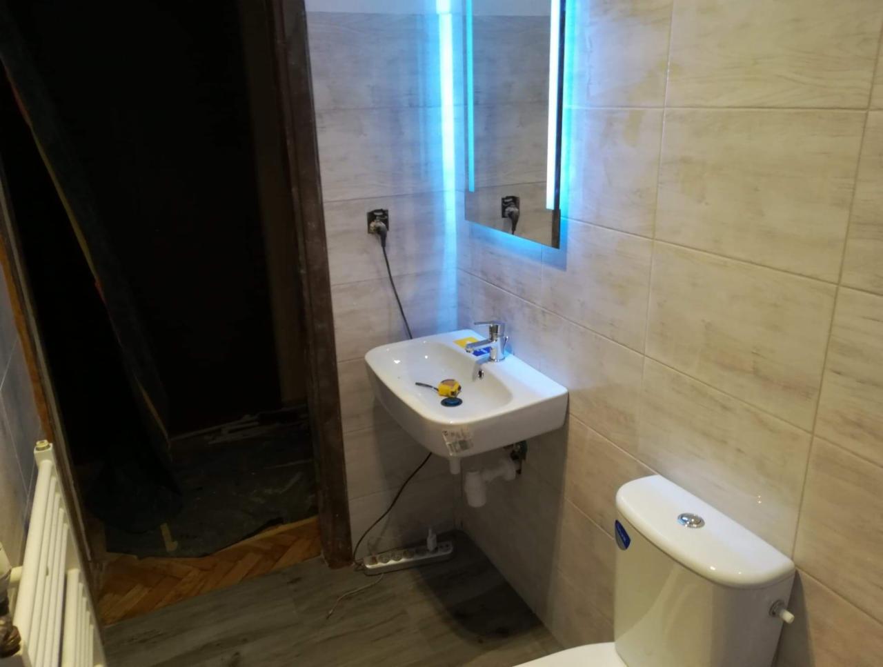 Nowa, jasna łazienka w trakcie remontu: umywalka podświetlana LED, biała toaleta i płytki imitujące kamień, widoczne narzędzia przy umywalce i niezakończone prace wykończeniowe przy drzwiach.