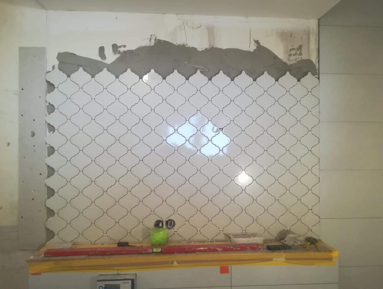 Montaż białych płytek arabeska na ścianie, widoczne ślady zaprawy, poziomica i inne narzędzia glazurnicze na dolnej krawędzi, etap prac wykończeniowych.