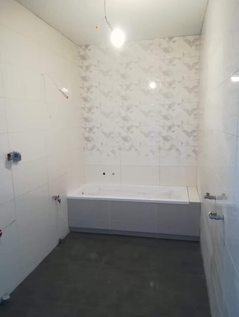 Wnętrze łazienki w trakcie wykańczania: białe płytki na ścianach, z dekoracyjnym wzorem na jednej ze ścian, zamontowana wanna obudowana płytkami, surowa podłoga, widoczne wystające przewody...