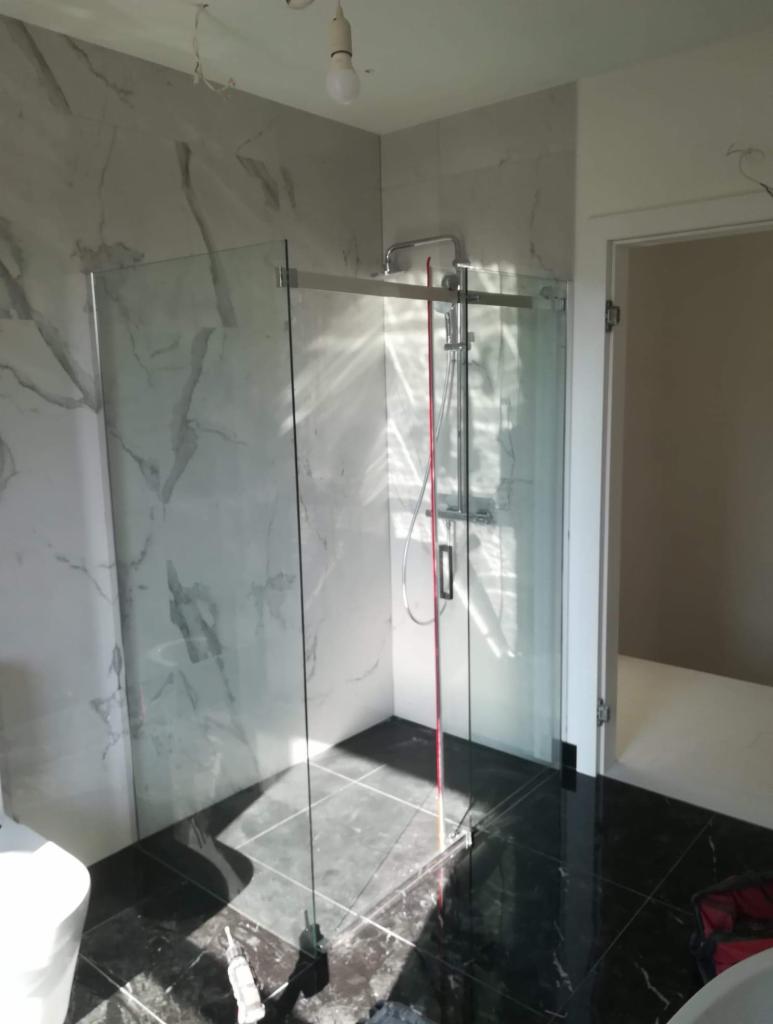 Nowoczesna kabina prysznicowa ze szklanymi ściankami, zamontowana w łazience z marmurowymi płytkami ściennymi i czarną, błyszczącą podłogą. Widoczny element instalacji prysznicowej i czerwony pręt...