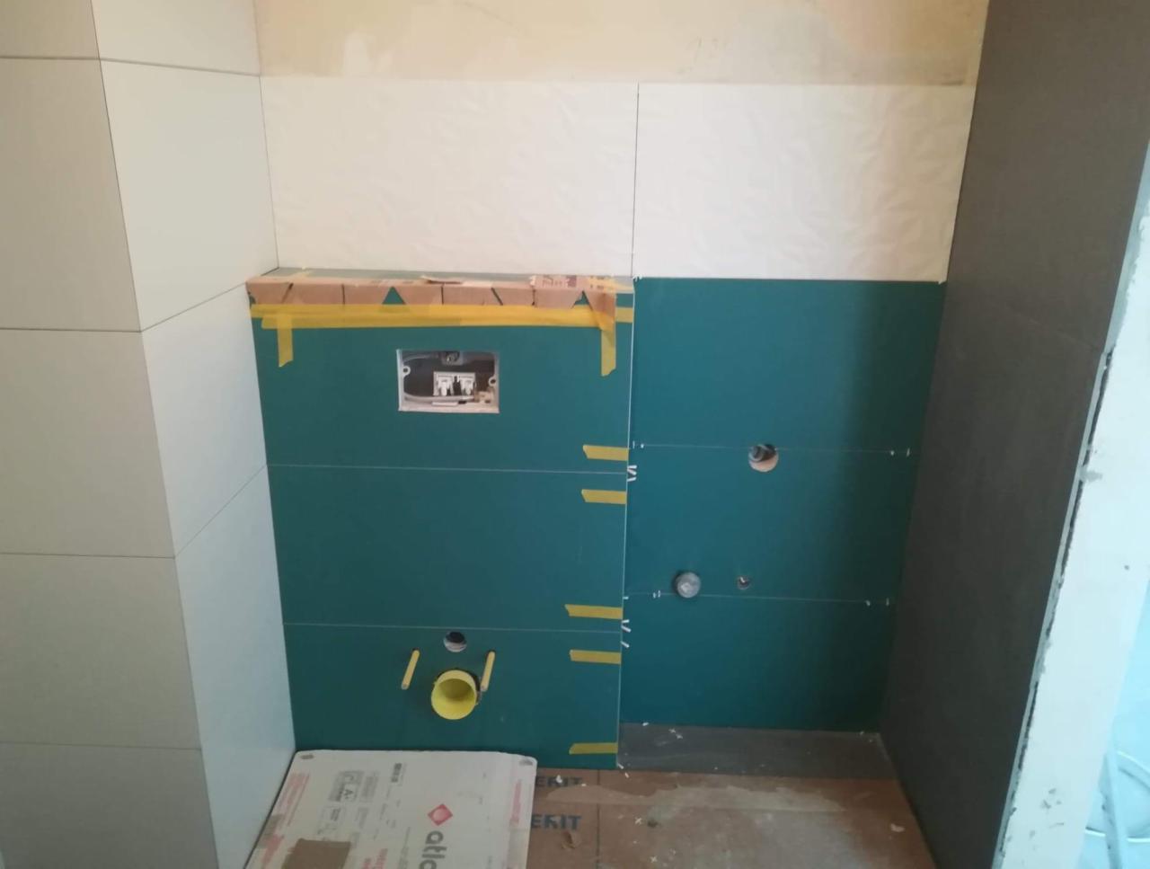 Przygotowanie ściany w łazience do układania płytek, widoczne zielone płyty kartonowo-gipsowe, taśma malarska, otwory na rury i puszka podtynkową WC, częściowo ułożone białe płytki na ścianie.