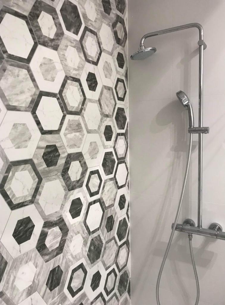 Ściana w łazience wyłożona heksagonalnymi płytkami w odcieniach szarości i bieli, z widoczną chromowaną baterią i słuchawką prysznicową.