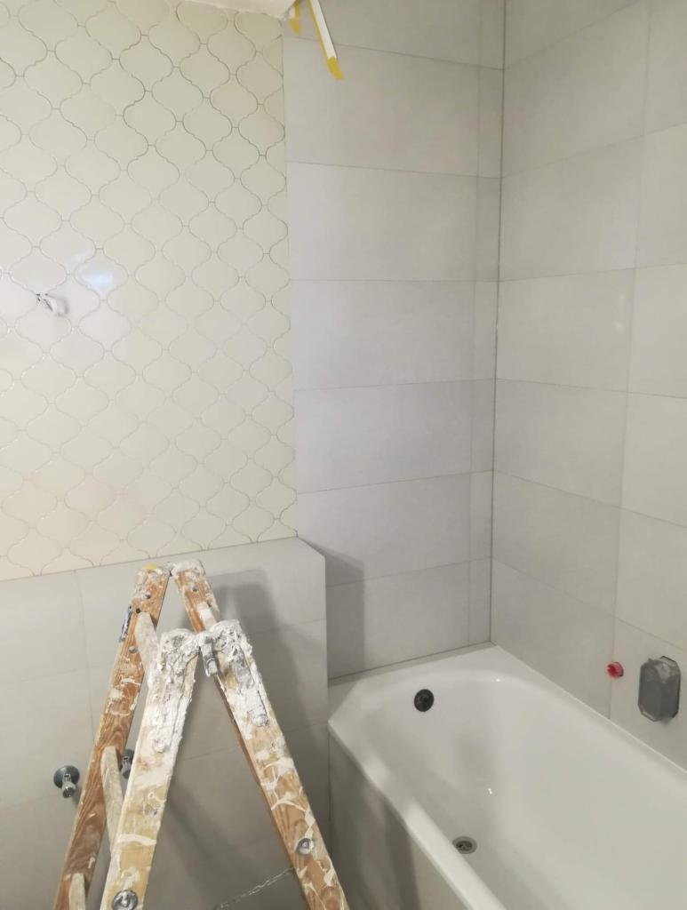 Biała łazienka w trakcie wykańczania: widoczna wanna, fragment ściany z położonymi białymi płytkami o wzorze marokańskim oraz drabina malarska z resztkami zaprawy.