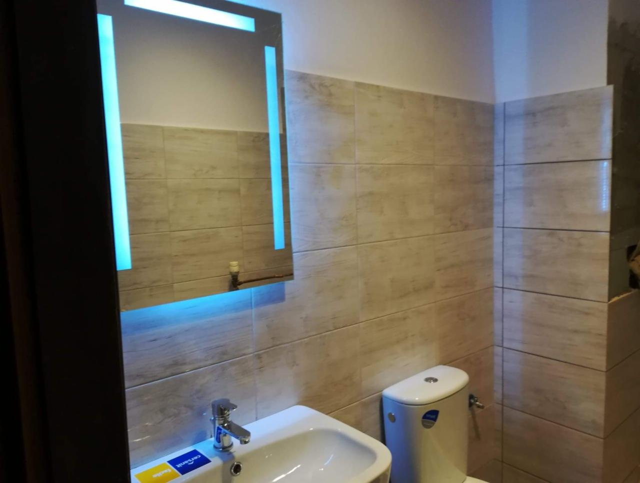 Wykończona łazienka w Warszawie z beżowymi płytkami imitującymi drewno, podświetlane lustro LED, biała umywalka i toaleta, widoczne detale armatury.