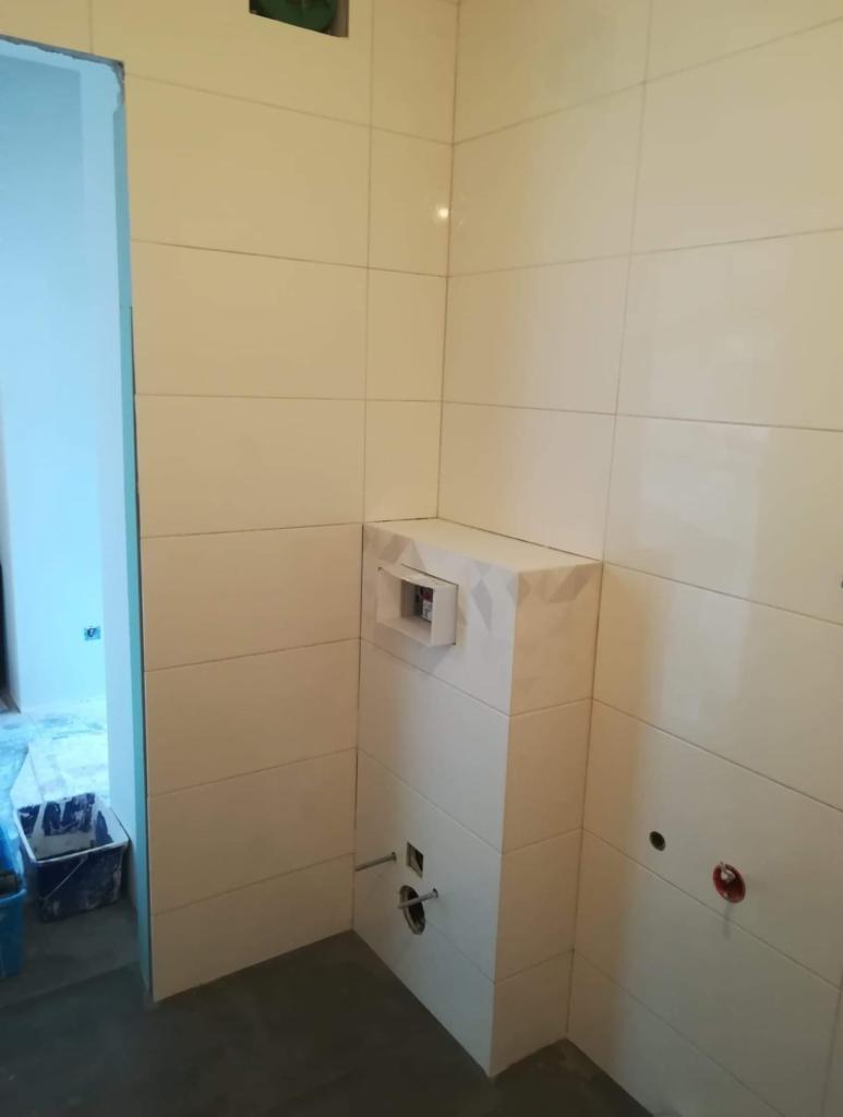 Świeżo wykafelkowana ściana w łazience z zabudową podtynkową WC, widoczne otwory na instalacje, ściany wyłożone jasnymi, prostokątnymi płytkami.
