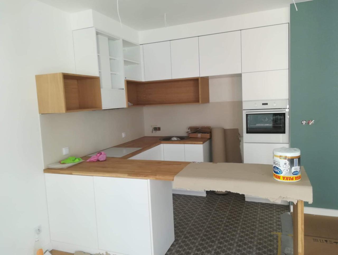 Wnętrze kuchni w trakcie remontu, z białymi szafkami, drewnianymi blatami, widocznym piekarnikiem i puszką farby na blacie roboczym, ściana pomalowana częściowo na zielono.