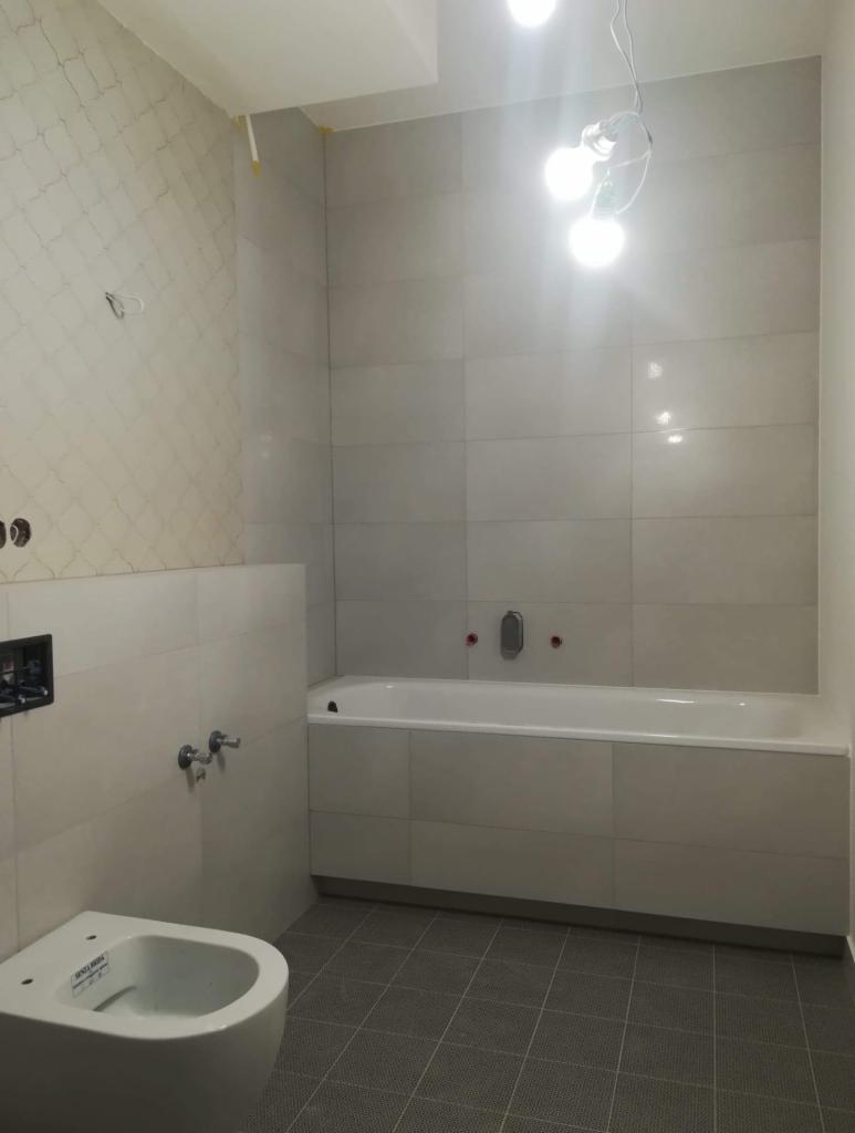 Wnętrze łazienki w trakcie wykańczania: biała wanna obudowana płytkami, podwieszany bidet, szare płytki podłogowe, niedokończone podłączenie oświetlenia sufitowego.