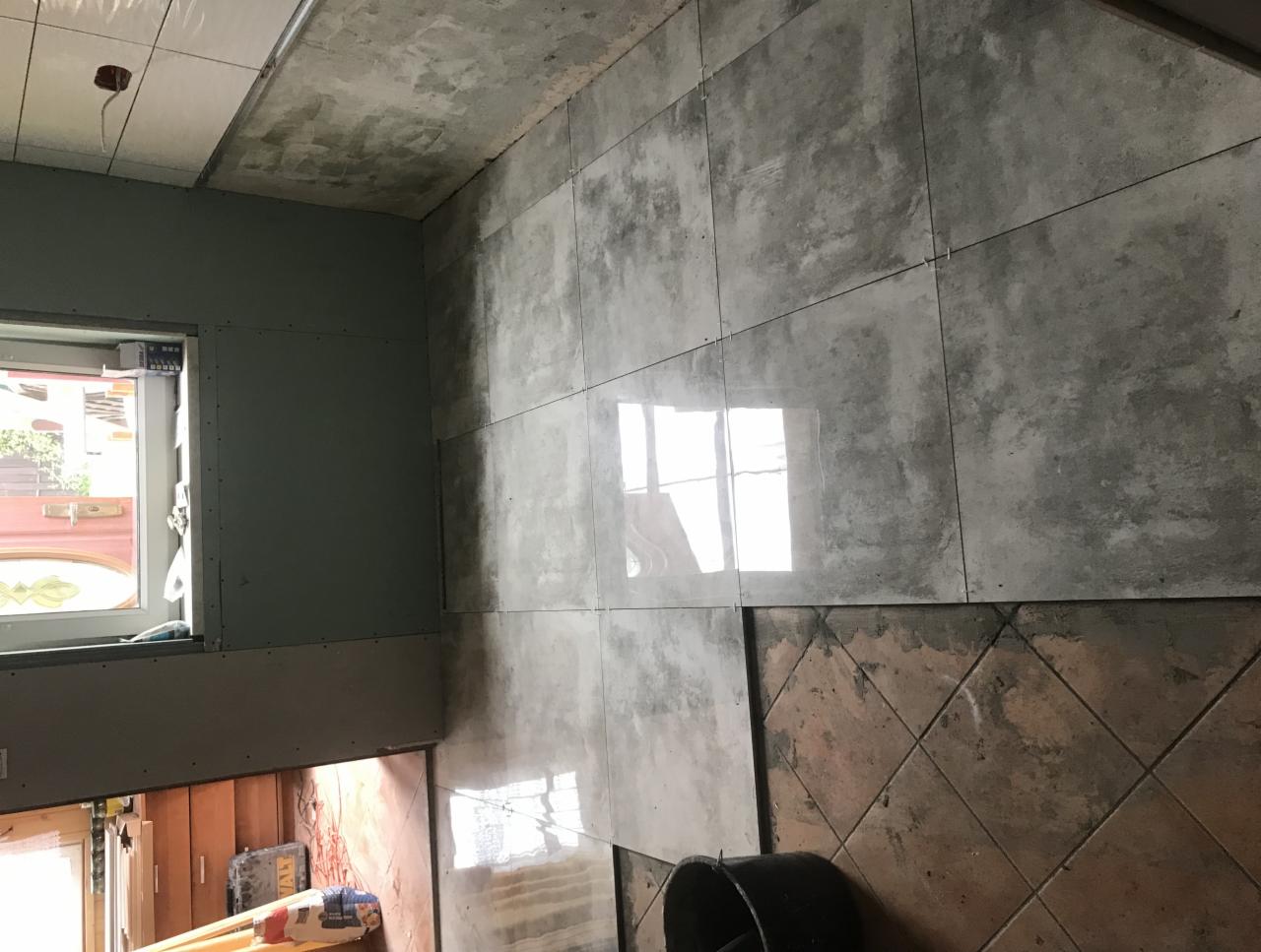 Montaż płytek ceramicznych na ścianie w łazience, widoczne szare płytki imitujące beton, częściowo ułożone, z widocznym odbiciem okna, sufit z płyt gipsowo-kartonowych, fragment starej podłogi...