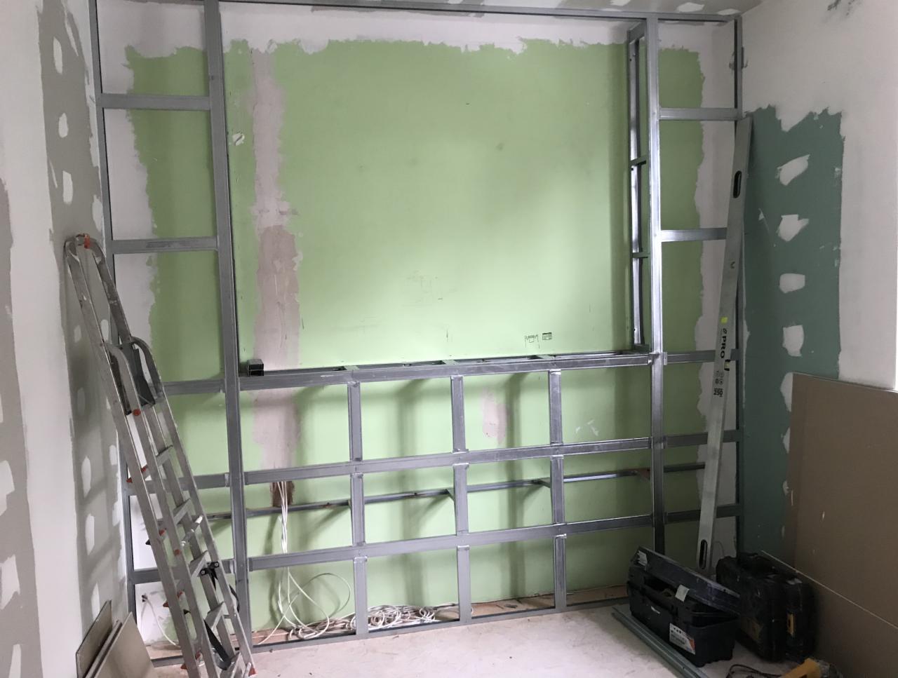 Konstrukcja metalowa pod zabudowę ściany z zielonym fragmentem ściany w tle, widoczna drabina, przewody elektryczne i skrzynki narzędziowe.