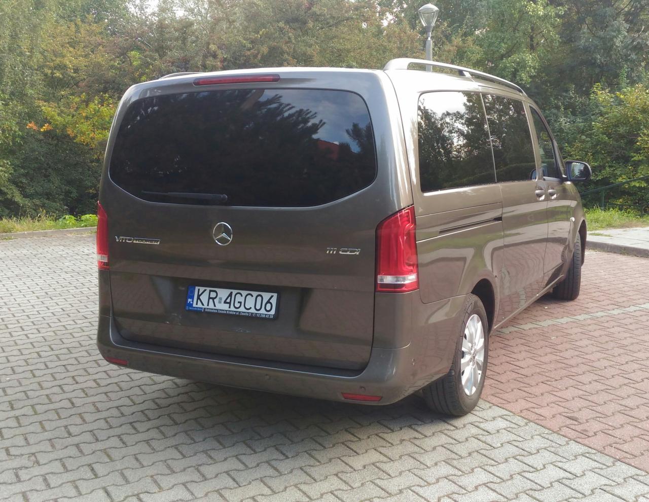 Szary Mercedes Vito Tourer 111 CDI z krakowską rejestracją KR-4GC06 zaparkowany na chodniku z kostki brukowej na tle zieleni.