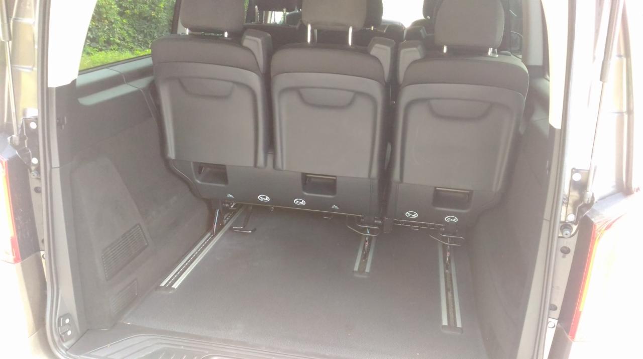 Wnętrze bagażnika busa z trzema rzędami siedzeń, widoczne szyny mocujące do podłogi, z tyłu zieleń.