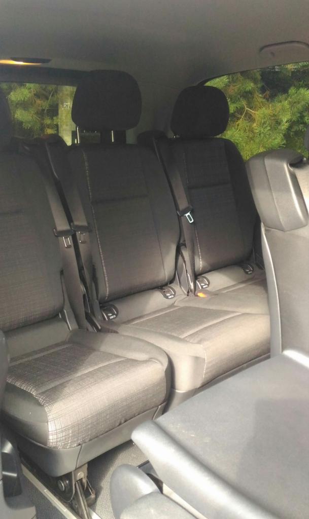 Wnętrze busa z trzema rzędami ciemnych foteli, widok z tyłu na zagłówki i pasy bezpieczeństwa, za oknem zieleń.