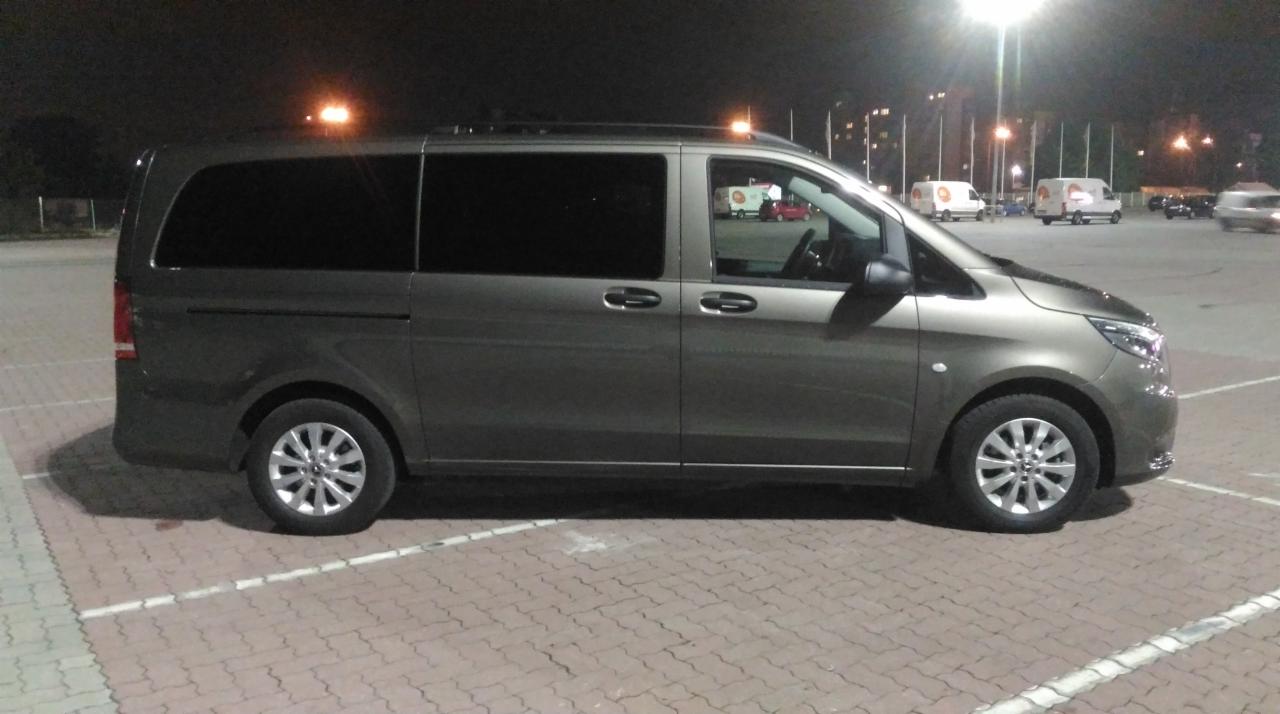 Szary van Mercedes-Benz V-klasa zaparkowany na parkingu z kostki brukowej w nocy, widoczne inne pojazdy w tle.