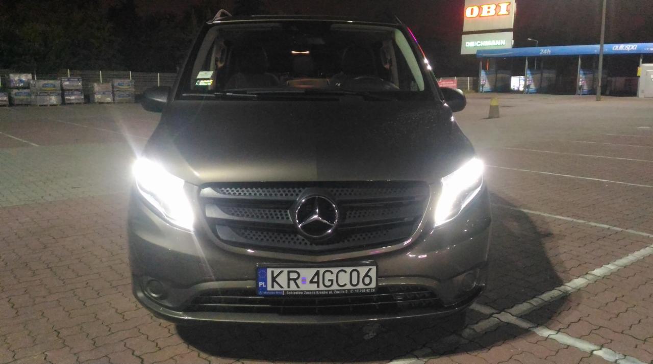 Przód brązowego samochodu marki Mercedes-Benz Vito z włączonymi światłami mijania na parkingu przed sklepem OBI nocą.