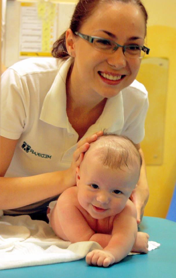 Uśmiechnięta kobieta w okularach, w białym uniformie z logo, delikatnie masuje główkę uśmiechniętego niemowlęcia leżącego na brzuszku na niebieskim stole przykrytym białą pieluszką.