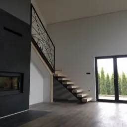 Nowoczesne schody z drewnianymi stopniami i geometryczną, czarną balustradą w przestronnym wnętrzu z kominkiem i dużym oknem tarasowym.