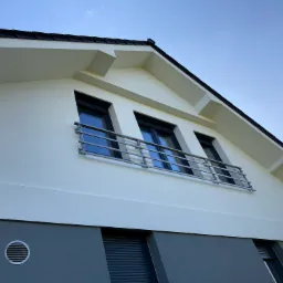 Biała fasada domu z trzema oknami zabezpieczonymi metalową balustradą balkonową, widok z dołu na tle błękitnego nieba.