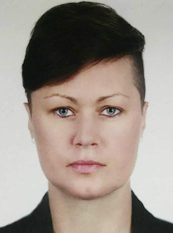 Portret kobiety z krótkimi, ciemnymi włosami i niebieskimi oczami, ubrana w ciemną marynarkę, na jasnym tle. Wyraz twarzy neutralny, oświetlenie równomierne.