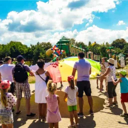 Grupa dzieci i dorosłych bawi się kolorową chustą animacyjną na tle placu zabaw i zieleni w słoneczny dzień.