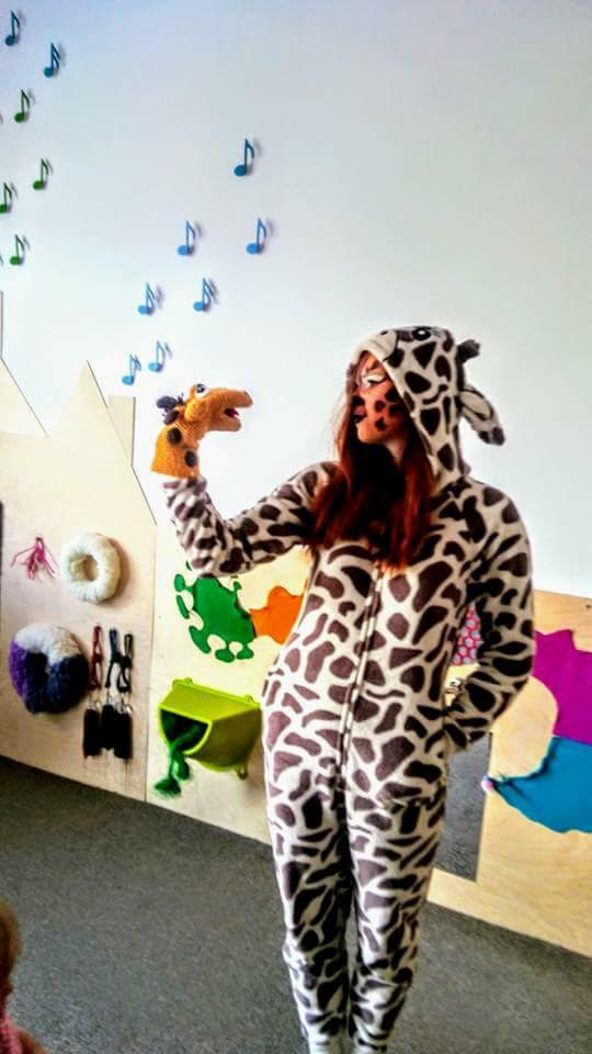Animator w kostiumie żyrafy prezentuje pacynkę żyrafy na tle ściany z dekoracjami muzycznymi i elementami wyposażenia sali zabaw.