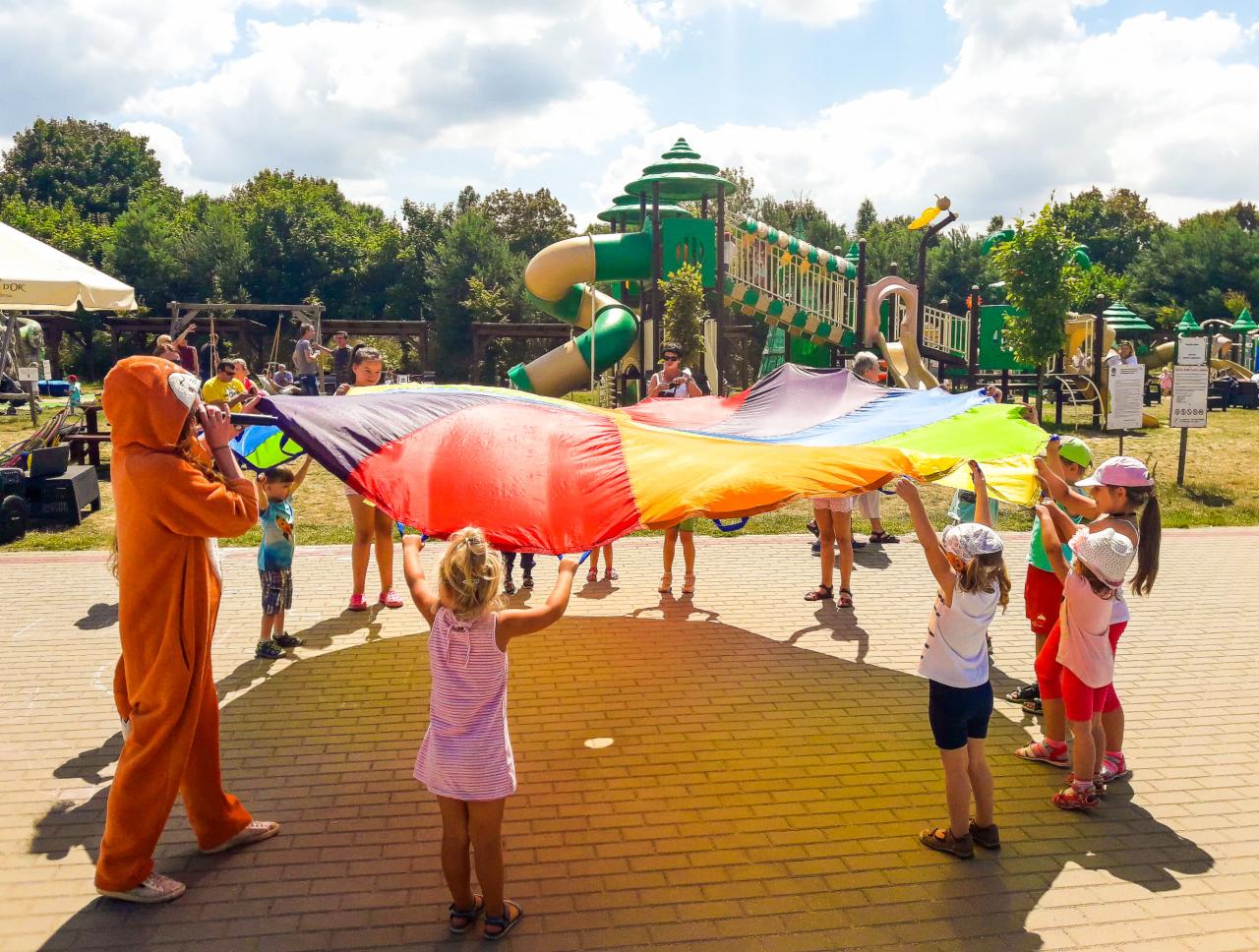 Grupa dzieci trzymająca kolorową chustę animacyjną na tle placu zabaw, animator w stroju zwierzęcia z boku.