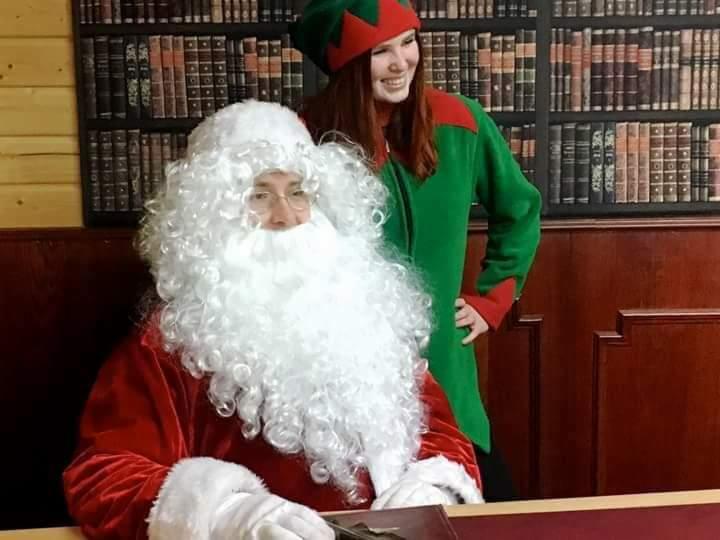 Mikołaj siedzący za biurkiem, piszący piórem, z uśmiechniętą elfką w zielonym stroju w tle, na tle regału z książkami.